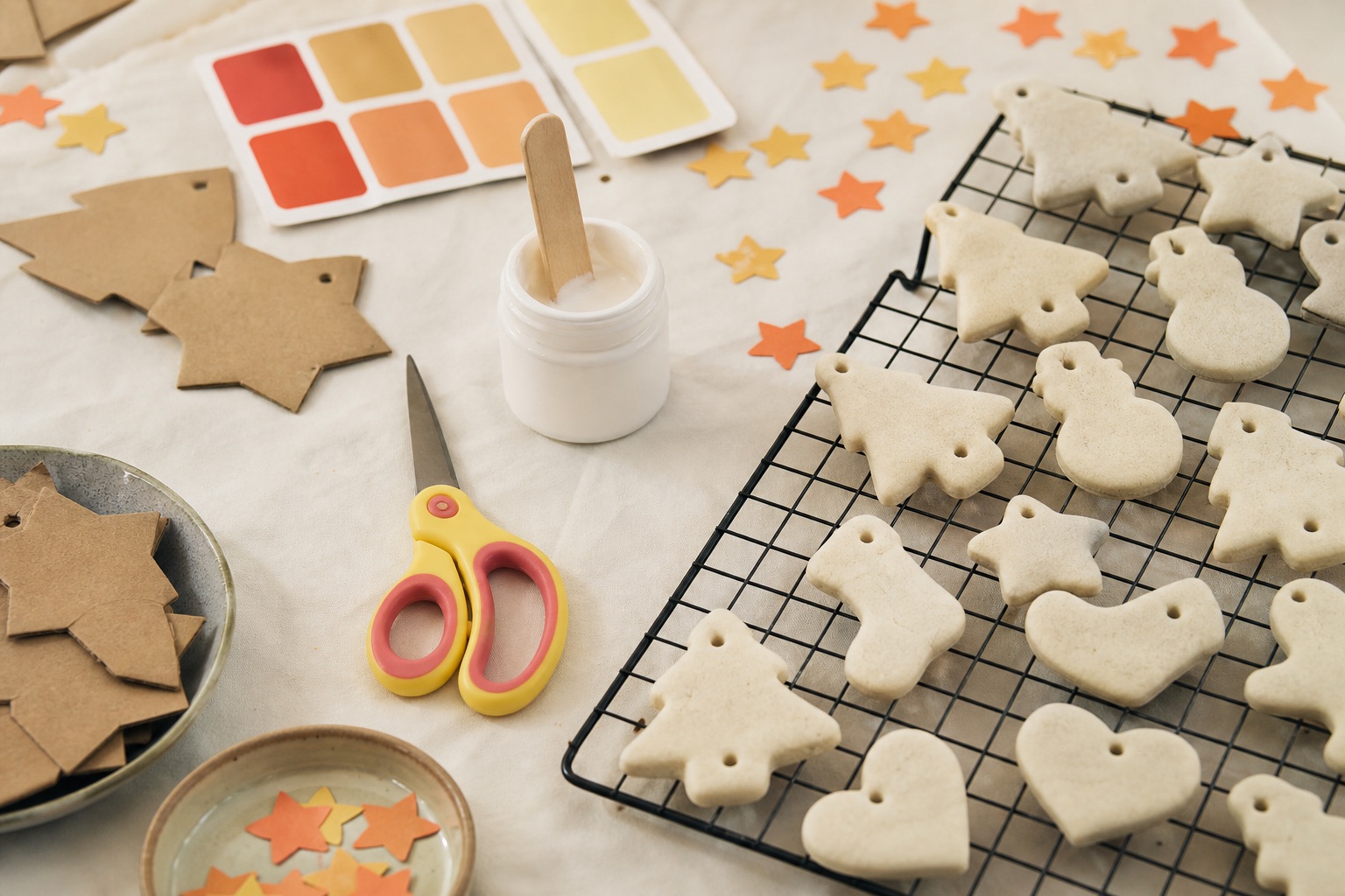 Overhead view of a kids craft table with salt dough ornaments drying on a rack, cardboard shapes, safety scissors and glue